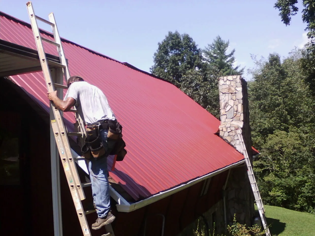 Roofer on a ladder beside a red metal roof and stone chimney — Soffit & Fascia Repair by Roofing Brothers in Barkhamsted